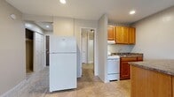 This single-floor unit offers an efficiently organized layout across a compact footprint. The living area serves as the central space, finished with neutral-toned carpet and textured ceilings. A sliding glass door dominates one wall, allowing natural light to fill the room while providing direct access to a private outdoor area enclosed by a wooden fence. A wall-mounted heating and cooling unit ensures year-round climate control.

The kitchen is practical and well-equipped, featuring warm oak cabinetry, dark granite-style countertops, and white appliances including a refrigerator and range with overhead ventilation. A peninsula extends into the space, providing additional counter surface and creating a natural division between the kitchen and living areas. The flooring transitions from tile in the kitchen to carpet in the adjacent rooms.

The bathroom is finished with matching tile flooring and features an oak vanity with granite-style countertops, a large mirror, and a glass-enclosed walk-in shower. The hallway connects the main living spaces and includes closet storage. Throughout the unit, neutral wall tones and consistent finishes contribute to a cohesive and functional aesthetic.