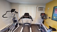 This single-floor fitness center offers a practical and functional space designed to support a well-rounded workout routine. The room features bold yellow accent walls that lend the space an energetic atmosphere, complemented by framed workout guides and informational posters that provide exercise references for users. The textured ceiling and neutral-toned walls in the cardio area create a no-frills, focused environment. Cardio equipment is a highlight of the space, with multiple treadmills, an elliptical trainer, and a stationary bike arranged to allow comfortable use. A cable strength training machine is also present, expanding the facility's versatility. A window fitted with vertical blinds allows for natural light and ventilation, and a wall-mounted air conditioning unit helps maintain a comfortable temperature during workouts. Dark wood-style flooring grounds the space and is easy to maintain. Informational charts on the walls guide users through exercise routines, adding an educational element to the facility. Overall, this fitness center is a well-equipped, purposefully arranged space suited for both cardio and strength training sessions.