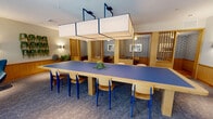 Located in Stamford, CT, this single-floor space offers a well-appointed and versatile layout designed for both collaboration and focused work. The entrance area welcomes visitors with an abundance of natural light streaming through large floor-to-ceiling glass doors and windows, complemented by lush potted plants, a sculptural concrete column, and a cozy seating arrangement featuring a modern blue accent chair and round side table.

The central conference area is anchored by a large, blue-topped communal table surrounded by light wood chairs with blue accents, creating a cohesive and contemporary aesthetic. Overhead, a striking arrangement of rectangular pendant light fixtures suspended on a blue metal frame provides focused, even illumination throughout the space.

Adjacent to the main table, a series of semi-private booth seating areas are framed by decorative wood-slatted partitions and patterned wallcoverings, offering intimate settings for smaller group meetings or individual work. Wall-mounted sconces provide warm, ambient lighting throughout.

A separate lounge zone features comfortable lounge chairs, a marble-top coffee table, and an impressive collection of tropical plants positioned against the floor-to-ceiling glazed facade, offering views of the surrounding greenery. The continuous blue-toned carpet ties all zones together, creating a cohesive and inviting atmosphere throughout this dynamic, professionally designed space.