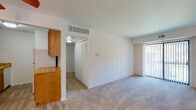 This single-floor unit offers a functional and straightforward layout across its main living spaces. The living area is generously sized, featuring wall-to-wall carpeting that adds warmth underfoot, crisp white walls, and a large sliding glass door fitted with vertical blinds that allows natural light to filter in and provides access to an outdoor area. A centrally located HVAC vent on the ceiling ensures efficient climate control throughout. Adjacent to the living space is a hallway area with a set of bi-fold closet doors, offering convenient built-in storage.

The kitchen is designed in a galley configuration, lined with honey-toned wood cabinetry on both upper and lower levels. It is equipped with a gas range, a full-size refrigerator, and a single-basin sink, all set against light wood-look vinyl plank flooring that extends through the kitchen and into the adjacent areas. The countertops complement the warm wood tones of the cabinetry.

The bathroom features a wood-framed vanity with ample drawer and cabinet storage, a white countertop, and a large mirror with overhead strip lighting. The wood-look flooring continues here, tying the space together with a cohesive aesthetic. Overall, the unit presents a clean, neutral palette that provides a versatile canvas for personalization.