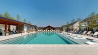 Located at 901 E Lincoln Hwy in Exton, Pennsylvania, this thoughtfully designed outdoor amenity space offers a resort-inspired experience centered around a generously sized swimming pool with crystal-clear turquoise water. The pool deck is lined with rows of white chaise lounges, providing ample space for sunbathing and relaxation beneath open skies. A striking covered pavilion with a prominent gabled roofline and rich wood framing serves as the architectural focal point, offering shaded seating that overlooks the pool area.

The surrounding architecture blends natural stone accents, wood detailing, and contemporary siding, creating a warm yet modern aesthetic. A covered lower terrace features stone columns, decorative planters with lush greenery and seasonal flowers, and lounge seating, connecting seamlessly to the pool deck via broad stone steps.

Beyond the pool, a separate outdoor social area provides grilling stations, dining tables and chairs, and a covered pergola — ideal for community gatherings. Adirondack chairs and fire pit elements offer additional relaxed seating areas along the paved walkway. Thoughtful landscaping with stacked stone retaining walls, young trees, and native plantings ties the entire space together, delivering a cohesive and inviting outdoor environment throughout the community.