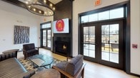 Located at 2060 Westgate Dr in Bethlehem, PA, this single-floor amenity space is designed with a contemporary industrial aesthetic, blending bold design elements with functional, community-focused areas. The welcoming entrance features a dramatic floor-to-ceiling black-framed glass partition that frames a private leasing or office area, set beneath a statement chandelier and complemented by large-format tile flooring. The main lounge area is anchored by a sleek fireplace clad in dark brick tile, with a wall-mounted TV above and generous seating arrangements bathed in natural light from expansive black-framed windows and French doors opening to an outdoor terrace. A fully equipped kitchenette serves the social space, offering dark cabinetry, a beverage cooler, microwave, and industrial-pipe open shelving adorned with curated décor. A dining nook with rustic horizontal wood-plank accent walls and Edison bulb pendant lighting provides a warm, inviting gathering spot. A dedicated game area features a classic shuffleboard table alongside modern seating and an additional wall-mounted TV. The fitness center is well-appointed with treadmills, ellipticals, spin bikes, free weights, and mirrors, all beneath exposed ductwork that reinforces the space's industrial character.