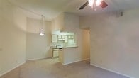 This single-story space features an open-concept layout that connects the living area and kitchen, creating a cohesive and functional flow throughout. The living area is finished with wall-to-wall carpeting in a neutral tone, contributing to a warm and understated atmosphere. A ceiling fan with an attached light fixture is mounted overhead, providing both air circulation and ambient lighting.

The kitchen is positioned centrally and defined by a raised pass-through counter that separates it from the living space while maintaining visual openness. White cabinetry lines the upper and lower walls of the kitchen, offering ample storage, and a range hood is installed above the stovetop. White appliances, including a stove and refrigerator, are integrated into the kitchen layout. A pendant light hangs near the kitchen edge, adding a focused lighting element to the dining or transitional area.

The walls throughout are finished in a light, neutral tone, lending a clean and bright feel to the interior. Vaulted or elevated ceiling height in the living area adds a sense of spaciousness, making the overall environment feel more open than its footprint might suggest. An arched or open doorway visible on one side provides passage to an additional area of the space.