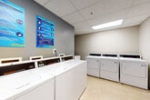 The shared laundry facility at Calibre Lake Apartments in Smyrna, Georgia, is a well-organized, single-floor space designed to serve the needs of residents efficiently. The room features a drop ceiling with recessed fluorescent panel lighting, providing consistent, bright illumination throughout. Light-toned vinyl tile flooring runs across the entire space, lending a clean and practical aesthetic.

The facility is equipped with a row of commercial-grade top-loading washers along one wall, accompanied by a matching set of front-loading dryers positioned opposite. Each machine features a digital control panel, and the layout allows for easy navigation between units. A folding table is available to residents for convenient garment handling after cycles are complete.

A water cooler is stationed in the corner of the room, adding a practical amenity for residents during longer laundry sessions. Informational and safety notices are displayed on the gray accent wall, keeping residents informed. The room is accessed through two gray doors — one solid panel door and one louvered door — the latter allowing for ventilation. The overall atmosphere is functional, tidy, and well-maintained, reflecting a commitment to resident convenience.