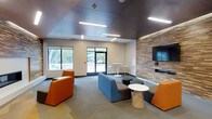 This single-floor communal lounge space is designed with a contemporary aesthetic that balances functionality and style. The room features a striking stacked stone accent wall serving as a focal point, adorned with a large mounted flat-screen television and a built-in ledge beneath. Sleek, dark ceiling panels are fitted with recessed linear LED lighting that evenly illuminates the space, contributing to a clean and modern atmosphere.

The lounge area is furnished with a mix of vibrant orange armchairs and a blue sectional sofa, complemented by round side tables and small circular ottomans in neutral tones. High-top bistro tables with white bar stools near the windows provide an alternative seating arrangement, making the space well-suited for both casual socializing and informal meetings.

Large windows and a glass-paneled door flood the interior with natural light and offer clear views of the outdoor surroundings, including what appears to be a covered pavilion and green landscaping. The neutral carpet flooring ties the varied furnishings together cohesively. Overall, this space presents a welcoming, energetic environment ideal for gathering, collaboration, or relaxation.