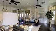 This single-floor space exudes a polished, contemporary character designed to impress and accommodate. The open-concept layout connects multiple lounge and reception areas seamlessly, creating a fluid sense of movement throughout. Dark hardwood flooring grounds the space with warmth, while crisp white walls and recessed lighting contribute to a bright, airy atmosphere. Multiple ceiling fans with dark finishes are suspended overhead, adding both a functional and stylish element to the design. The main seating area features sleek charcoal sofas accented with vibrant blue, teal, green, and yellow throw pillows, bringing a lively energy to the interior. A round wooden coffee table serves as a focal point, complemented by elegant table lamps that add a soft, inviting glow. In the background, a glazed reception or service counter is visible, suggesting a professional leasing or community office function. Arched doorways and open passageways connect to additional lounge areas, one of which is visible with access to a glass-paneled exterior door that floods the space with natural light. The overall ambiance strikes a balance between sophisticated design and comfortable livability, making it an ideal gathering or welcoming environment.