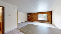 This single-floor unit offers a straightforward and functional layout centered around an open-concept living and kitchen area. The living room is a standout space, distinguished by a bold diagonal wood-paneled accent wall in warm, natural tones that adds character and visual interest to the room. Carpeted flooring runs throughout the main living area, while tile flooring transitions into the kitchen zone. A window on the wood-paneled wall allows natural light to filter in and provides views of the surrounding outdoors. The space is equipped with a wall-mounted air conditioning unit and a baseboard heater, offering year-round climate control. A ceiling fan with a built-in light fixture serves the kitchen and dining area, which features a peninsula counter with a dark laminate surface and wood cabinetry — offering both workspace and a casual bar-style seating option. Upper cabinets and an under-cabinet light complete the kitchen area. The bathroom includes a full tub-and-shower combination with classic white square tile surround, a toilet, and tile flooring, presenting a clean and utilitarian design. Overall, the space offers a practical and livable floor plan with a retro aesthetic rooted in its wood paneling and neutral palette.