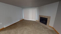 Unit A103 is a well-appointed single-floor residence offering a practical and comfortable layout. The living room serves as the heart of the unit, featuring a tile-surround fireplace as a standout focal point, plush carpeting, and a large sliding glass door with vertical blinds that allows natural light to fill the space. The transition from the living area to the rest of the unit is marked by rich, dark hardwood-style flooring that extends throughout the kitchen, hallway, and entry areas.

The kitchen is efficiently designed with warm honey-toned cabinetry, grey laminate countertops, and a full suite of white appliances including a coil-top electric range, refrigerator, and dishwasher. A ceiling fan is present in the open area connecting the kitchen and living space, improving air circulation throughout.

The hallway leads to a bedroom featuring soft carpeting, a window with a view of surrounding greenery, and baseboard heating. The full bathroom is finished with a warm wood-toned vanity, grey countertop, wall-mounted lighting above the mirror, and a full-size tub and shower combination set against clean white surroundings. The unit also includes closet storage and baseboard heating throughout, rounding out a functional and well-organized living space.