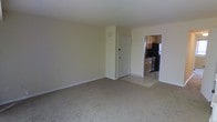 This two-floor residential space offers a straightforward and functional layout suited for everyday living. The main living area is finished with wall-to-wall carpeting, providing a warm and comfortable underfoot feel, while neutral-toned walls create a clean, versatile backdrop that accommodates a variety of interior styles. The room is generously proportioned, offering ample space for living and dining arrangements. Adjacent to the living area, the kitchen is accessible through an open doorway and features wooden cabinetry with a warm brown finish, providing substantial storage space. The kitchen is equipped with dark appliances, including a refrigerator and a range, and transitions seamlessly to tile flooring, offering a practical and easy-to-maintain surface. A white interior door near the kitchen entry adds a clean contrast to the surrounding neutral palette. White baseboards run throughout the visible areas, contributing to a tidy and cohesive finish. The space spans two floors, suggesting additional rooms and living areas beyond what is immediately visible. Overall, the layout prioritizes practicality and livability, making it a solid and functional residential option.