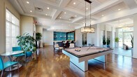 This expansive single-floor amenity space is designed with a contemporary aesthetic, combining functional gathering areas with upscale finishes throughout. Rich hardwood flooring runs continuously across the open layout, complementing the neutral gray and white color palette accented by bold teal details.

The main lounge area features a tufted sectional sofa anchored by a geometric area rug, a low-profile coffee table, and floor-to-ceiling windows with views to the exterior, flooding the space with natural light. Large glass entry doors open directly to the outdoors, enhancing the sense of connectivity.

A dedicated billiards area is centered around a sleek white pool table beneath a statement pendant light fixture, with a coffered ceiling above adding architectural depth. Adjacent to the game area, a curved wet bar with dark cabinetry and a sweeping white quartz countertop serves as the social hub, accented by a vibrant teal subway tile backsplash and under-counter beverage refrigerators.

A separate lounge nook features a linear gas fireplace set into a stacked stone accent wall, built-in display shelving, and a wall-mounted television. A private meeting or sitting room, accessible via a decorative sliding partition door, offers teal accent chairs and patterned carpeting. Lush potted greenery is placed throughout, adding warmth to the polished, modern atmosphere.