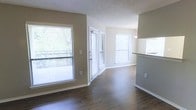 This single-floor residential space offers a clean, functional layout highlighted by a cohesive neutral color palette and rich, dark hardwood-style flooring that runs throughout the main living area. The open living space is bathed in natural light, courtesy of multiple large windows fitted with horizontal blinds, as well as a full glass-paneled door that leads to an outdoor balcony or patio. The textured popcorn ceiling adds a classic finish overhead, while crisp white baseboards and trim provide a sharp contrast against the warm taupe-toned walls. The living area flows seamlessly toward a partial kitchen enclosure, where a raised countertop with a dark surface is visible, suggesting a breakfast bar or pass-through counter. A pendant light fixture is subtly visible above this area, adding a touch of functional style. The glass door and surrounding windows allow generous views of mature trees outside, bringing a sense of greenery and the outdoors into the space. Overall, the unit presents a well-lit, move-in-ready environment with practical finishes suited for comfortable everyday living.
