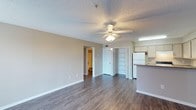 This single-floor unit features an open-concept layout that seamlessly connects the living area and kitchen, creating a functional and cohesive living space. The living area is finished with dark hardwood-style flooring that extends throughout, providing a clean, modern contrast against the neutral beige walls and white baseboards. A ceiling fan with an integrated light fixture is centrally positioned overhead, offering both ventilation and ambient lighting. The textured ceiling adds a classic architectural detail consistent throughout the space.

A built-in shelving unit with multiple tiers is situated near the center of the unit, offering practical open storage and a subtle design element. An interior doorway provides direct access to a bathroom, which is partially visible and features standard fixtures. Adjacent to the living area, the kitchen is defined by a raised peninsula counter that serves as a natural divider. White cabinetry lines the upper walls, complemented by a white refrigerator and under-cabinet fluorescent lighting. The overall layout prioritizes practicality, with clear sightlines across the space and straightforward access to all areas.