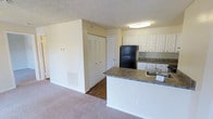This single-floor residential unit offers a practical and well-organized layout designed for comfortable everyday living. The open-concept design seamlessly connects the kitchen and living area, creating a cohesive and functional space. The kitchen is equipped with white cabinetry offering generous storage, dark granite-style countertops, a black refrigerator, and a built-in sink. A peninsula counter extends into the living space, providing additional workspace and a natural boundary between the kitchen and the carpeted living area. The living area features soft, neutral-toned carpet and benefits from a floor-level HVAC vent, ensuring consistent climate control throughout the space. A textured popcorn ceiling adds a classic residential character overhead, while flush-mounted ceiling lighting provides even illumination. A hallway off the main living area leads to additional rooms and features a large double-door closet with louvered bi-fold doors, offering substantial storage capacity. The overall color palette is light and neutral, with white walls and trim contributing to a clean, airy ambiance throughout the unit.