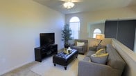 This two-story home showcases a thoughtfully designed living space that combines comfort and style. The main living area is highlighted by soaring ceilings that create an airy, open atmosphere, complemented by a ceiling fan that enhances the room's functionality. Distinctive arched windows are a standout architectural feature, allowing generous amounts of natural light to fill the space while adding a refined, classic touch to the interior.

The living room is well-appointed with neutral-toned carpet and light-colored walls that create a clean, versatile backdrop. The seating arrangement centers around a tufted leather ottoman that doubles as a coffee table, flanked by coordinating gray upholstered chairs and a sofa accented with yellow and white throw pillows. A sleek black media console provides dedicated space for entertainment needs. A tall indoor plant introduces a natural element, adding life and texture to the corner of the room. A secondary arched window is visible in the adjacent area, reinforcing the home's consistent architectural detailing and ensuring the space remains bright and inviting throughout.