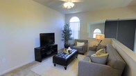 This two-story home showcases a thoughtfully designed living space that combines comfort and style. The main living area is highlighted by soaring ceilings that create an airy, open atmosphere, complemented by a ceiling fan that enhances the room's functionality. Distinctive arched windows are a standout architectural feature, allowing generous amounts of natural light to fill the space while adding a refined, classic touch to the interior.

The living room is well-appointed with neutral-toned carpet and light-colored walls that create a clean, versatile backdrop. The seating arrangement centers around a tufted leather ottoman that doubles as a coffee table, flanked by coordinating gray upholstered chairs and a sofa accented with yellow and white throw pillows. A sleek black media console provides dedicated space for entertainment needs. A tall indoor plant introduces a natural element, adding life and texture to the corner of the room. A secondary arched window is visible in the adjacent area, reinforcing the home's consistent architectural detailing and ensuring the space remains bright and inviting throughout.