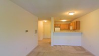This single-floor residential unit offers a practical and comfortable open-concept layout designed for everyday living. The living area features neutral wall-to-wall carpeting that lends a warm, cohesive feel to the space, while flush-mount ceiling lighting provides consistent illumination throughout.

The kitchen is positioned adjacent to the main living area and is defined by a raised breakfast bar counter, which serves both as a functional workspace and a natural divider between the two zones. Warm honey-toned wood cabinetry lines the upper and lower walls of the kitchen, offering generous storage. A white refrigerator and additional white appliances are visible, contributing to a clean, uniform aesthetic.

A central structural column subtly delineates the living space from the entry corridor, which leads to a white paneled front door. A wall-mounted HVAC vent and thermostat panel are visible along the main wall, indicating a dedicated climate control system. The textured ceiling adds a subtle architectural detail consistent throughout the unit.

The layout flows efficiently from the entry through the living area and into the kitchen, making the space feel connected and easy to navigate. Overall, the unit presents a straightforward, functional interior with a neutral palette well-suited for personalization.