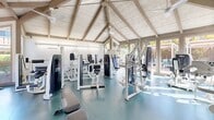 The Glenbrook Apartments Strength Center in Cupertino, CA is a dedicated fitness facility designed to support a comprehensive strength training experience. The space is housed within a striking pavilion-style structure featuring soaring vaulted ceilings with exposed wooden beam trusses and white-painted shiplap paneling, lending the room an open, airy, and inviting atmosphere.

Large windows line the walls on multiple sides, flooding the interior with abundant natural light and offering views of the surrounding greenery. Ceiling fans are mounted along the roofline, ensuring comfortable airflow throughout workouts. The floor is covered in durable teal-toned rubber gym flooring, providing both safety and functionality.

The facility is outfitted with a robust selection of equipment. A full lineup of Cybex and Eagle selectorized strength machines targets major muscle groups, including chest, back, shoulders, and legs. A multi-tiered dumbbell rack offers a wide range of free weight options, complemented by adjustable benches and a stability ball for versatile training. Additional cardio options, including a stationary bike and a rowing-style machine, round out the offerings. Workout reference charts are posted on the walls, providing guidance for users at all fitness levels. The overall ambiance is bright, clean, and well-maintained, making this strength center a standout amenity for residents.
