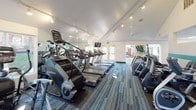 This fitness center is a spacious, single-floor facility designed to support a comprehensive workout experience. The main cardio area is lined with a generous row of treadmills, StairMaster climbers, and elliptical machines, all positioned to take advantage of large windows that flood the space with natural light. Mirrored walls extend the sense of openness while allowing users to monitor their form during exercise. Track lighting runs along the white ceiling, complementing the bright, energetic atmosphere throughout.

Adjacent to the cardio zone, a dedicated strength training section houses cable machines and multi-use weight equipment, offering a full range of resistance training options. A separate functional training room provides open floor space ideal for stretching, balance work, and group activity, equipped with stability balls, a wall-mounted television, and additional windows overlooking greenery outside.

The facility also includes a convenient refreshment and snack station stocked with beverages and supplements, as well as a water fountain. Ceiling fans and a well-ventilated layout contribute to a comfortable workout environment. Throughout, a multi-toned blue and grey carpet ties the space together visually, while teal accent details add a modern, cohesive design touch. The overall ambiance is clean, bright, and purpose-built for fitness.