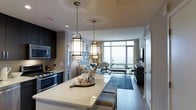 This single-floor residence showcases a thoughtfully designed open-concept layout that seamlessly connects the kitchen, dining, and living areas into one cohesive, flowing space. The kitchen is outfitted with dark cabinetry, stainless steel appliances including a built-in microwave and range, and a generous white quartz waterfall island that doubles as a breakfast bar with upholstered stools beneath. Two striking lantern-style pendant lights hang above the island, adding a sophisticated focal point to the space.

The dining area transitions naturally into the living room, which is anchored by a plush sofa, accent chairs, and a richly toned blue area rug atop hardwood flooring. A wall-mounted flat-screen television is positioned near a dark wood media console. Floor-to-ceiling windows along the far wall flood the living area with abundant natural light while offering expansive views beyond. Framed artwork adorns the walls, and layered lighting — including table lamps and recessed fixtures — contributes to the warm, polished ambiance throughout. The overall aesthetic is contemporary and refined, with a cohesive neutral palette accented by navy and steel blue tones.