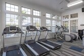Located at 29980 FM-2978 in Magnolia, Texas, this well-appointed fitness center offers a comprehensive workout environment designed to support a full range of exercise routines. The single-floor facility is generously sized, providing ample space for both cardio and strength training activities without feeling crowded.

The interior is bright and inviting, thanks to an abundance of large, multi-pane windows fitted with white shutters that line the walls and allow natural light to flow throughout the space. Ceiling fans are mounted overhead to ensure comfortable airflow during workouts, complemented by flush fluorescent lighting for consistent illumination.

Distinctive patterned carpeting in neutral tones with blue accents covers the floor, lending the space a modern, polished aesthetic. Full-length mirrors span an entire wall, visually expanding the room and allowing users to monitor their form during exercise.

The equipment selection is extensive and well-maintained, including multiple treadmills, cable and selectorized strength machines, a multi-rack dumbbell station with a full range of free weights, leg press and extension machines, and upright cycling equipment. Wall-mounted televisions are positioned throughout for entertainment during workouts.

Crown molding details along the ceiling add an unexpected architectural elegance to the space. An exit door with glass panels provides additional outdoor visibility, contributing to the open, airy atmosphere of this fully equipped community fitness center.