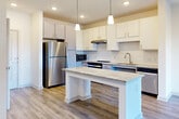 This well-designed single-floor unit offers a clean, contemporary aesthetic with consistent wood-look luxury vinyl plank flooring running throughout every room. The heart of the space is an open-concept kitchen that flows seamlessly into the living area. The kitchen is equipped with crisp white cabinetry, granite countertops, a white subway tile backsplash, and a full suite of stainless steel appliances including a refrigerator, range, microwave, and dishwasher. A central kitchen island with a matching granite surface provides additional prep space and casual seating potential. Pendant lights hang above the island, adding both task lighting and a stylish focal point. The adjacent living area benefits from natural light through a sliding glass door that opens to an outdoor view, complemented by a ceiling fan overhead. The bedrooms are bright and airy, each featuring windows with outdoor views and ceiling fans for year-round comfort. The bathroom showcases an accessibility-conscious design with a wide vanity featuring granite countertops, a large mirror, and grab bar support. A dedicated in-unit laundry room houses a full-size front-loading washer and dryer. Throughout, the neutral color palette and clean finishes create a bright, functional, and inviting living environment.