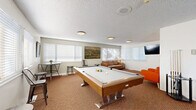 Located in Lakewood, Colorado, this single-floor clubhouse offers a well-rounded community gathering space designed for both recreation and socializing. The layout encompasses a combined living and game room, a dining room, and a kitchen, all finished with warm, textured carpeting throughout the main areas and light hardwood-style flooring in the kitchen.

The living and game room serves as the centerpiece of the space, featuring a full-size pool table as a standout recreational element, complemented by an air hockey table, a leather sofa, and vibrant accent seating. A wall-mounted flat-screen TV and landscape artwork add character to the room, while multiple windows dressed with white blinds allow ample natural light to fill the space.

The dining area is thoughtfully arranged with a large high-top communal table surrounded by industrial-style metal chairs, providing generous seating for group gatherings. Sliding room dividers offer flexible space configuration, and large vertical-blind windows bathe the room in natural light.

The kitchen is clean and modern, outfitted with gray shaker-style cabinetry, light quartz countertops, a stainless steel dishwasher, and a microwave. A window above the sink brings in additional natural light. The textured ceilings and flush-mount lighting fixtures are consistent throughout, giving the entire clubhouse a cohesive, well-maintained appearance.