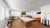 Located in Lakewood, Colorado, this single-floor clubhouse offers a well-rounded community gathering space designed for both recreation and socializing. The layout encompasses a combined living and game room, a dining room, and a kitchen, all finished with warm, textured carpeting throughout the main areas and light hardwood-style flooring in the kitchen.

The living and game room serves as the centerpiece of the space, featuring a full-size pool table as a standout recreational element, complemented by an air hockey table, a leather sofa, and vibrant accent seating. A wall-mounted flat-screen TV and landscape artwork add character to the room, while multiple windows dressed with white blinds allow ample natural light to fill the space.

The dining area is thoughtfully arranged with a large high-top communal table surrounded by industrial-style metal chairs, providing generous seating for group gatherings. Sliding room dividers offer flexible space configuration, and large vertical-blind windows bathe the room in natural light.

The kitchen is clean and modern, outfitted with gray shaker-style cabinetry, light quartz countertops, a stainless steel dishwasher, and a microwave. A window above the sink brings in additional natural light. The textured ceilings and flush-mount lighting fixtures are consistent throughout, giving the entire clubhouse a cohesive, well-maintained appearance.