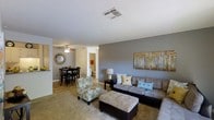 This single-floor residence offers a thoughtfully designed open-concept layout that seamlessly connects the living and dining areas, creating a cohesive and inviting atmosphere. The living area is anchored by a generously sized sectional sofa accented with decorative pillows in soft blues and neutrals, complemented by a tufted ottoman and a floral accent chair that add character and comfort. A nature-inspired canvas artwork decorates the bold gray feature wall, providing a striking visual focal point. The space is finished with plush wall-to-wall carpeting underfoot, contributing to a warm and cozy ambiance. Overhead, a textured ceiling with centrally located HVAC venting ensures year-round comfort. The adjacent dining area accommodates a dark-toned dining table and chairs, lit by a ceiling fan with integrated lighting, making it ideal for everyday meals or casual entertaining. A decorative sunburst mirror adds a stylish accent to the dining nook's wall. Beyond the main living space, a hallway leads to additional rooms, hinting at a well-organized and functional floor plan. The overall aesthetic balances contemporary design with homey warmth, offering a comfortable and practical single-level living experience.