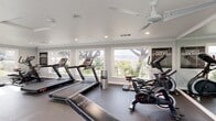 Located in Temecula, CA, this fitness center is a comprehensively equipped exercise facility designed to support a full range of workout routines. The space is bright and welcoming, featuring large windows along multiple walls that flood the interior with natural light while offering scenic views of the surrounding trees, greenery, and rolling hillside landscape — providing an inspiring backdrop for any workout.

The flooring throughout consists of durable, dark rubberized material well-suited for heavy equipment and high-traffic use. Recessed lighting and a ceiling fan ensure a comfortable environment, while crown molding and clean white ceilings lend a polished, well-maintained appearance.

The cardio zone is prominently featured, housing multiple treadmills and stationary spin bikes, along with an elliptical and stair-stepper machine. The strength training area is equally well-appointed, including a multi-function cable machine, a variety of weight benches, adjustable dumbbells on tiered racks, medicine balls, and additional resistance equipment. Full-length mirrors are positioned along one wall, enhancing the sense of space and supporting proper form during workouts.

Fitness center rules are clearly posted, and the layout allows for ample circulation between equipment. This facility offers everything needed for a thorough, effective workout in a bright, comfortable setting.