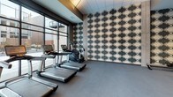 This contemporary fitness center is thoughtfully designed to support a full range of workout routines across a single, open floor. The space is anchored by exposed concrete columns that lend an industrial-chic aesthetic, complemented by large floor-to-ceiling windows and glass doors that flood the interior with natural light and offer views of an adjacent outdoor terrace. Large-format gray tile flooring extends throughout, providing a clean, durable surface underfoot.

The cardio zone is well-appointed with a row of treadmills, elliptical machines, stationary bikes, and a rowing machine, many equipped with digital interactive displays. A glass-enclosed studio area nearby provides a dedicated space for focused training, featuring framed artwork and a reflective glass wall. Strength training is equally well-represented, with a cable and multi-functional weight machine, adjustable benches, and a well-stocked free weights rack with dumbbells and exercise balls.

A bold geometric black-and-white feature wall adds a striking visual element, enhancing the modern ambiance of the space. Recessed and cove lighting in the ceiling creates a layered, polished look throughout. A private room with a wood-paneled door offers additional utility, likely serving as a restroom or changing area. The overall design balances functionality with refined aesthetics, creating an inviting environment for fitness.