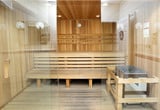 This single-level wellness space is thoughtfully designed to accommodate both relaxation and accessibility. The room is anchored by a walk-in, roll-accessible shower equipped with stainless steel grab bars, a fold-down shower bench, a rain showerhead, and a handheld spray — catering to a wide range of users with care and practicality.

The standout feature of the space is a generously sized, glass-enclosed cedar sauna. Warm-toned tongue-and-groove cedar lines the walls, benches, and ceiling, creating an inviting atmosphere. Recessed lighting and decorative etched glass panels add refined detail, while tiered benching offers ample seating for multiple occupants.

The surrounding wet room and corridor area feature light wood-look tile flooring that extends throughout, contributing to a cohesive and spa-like aesthetic. White textured tile wainscoting trimmed with a bold cobalt blue accent band lines the lower walls, adding a distinctive design touch. A decorative mirror is mounted on one wall, and multiple doorways provide convenient access throughout the space. The overall ambiance is clean, bright, and purpose-built for wellness and comfort.
