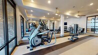 Located at 2421 Landmark Dr in Raleigh, NC, this expansive fitness center is a well-appointed, single-floor facility designed to support a comprehensive range of workout routines. The space is thoughtfully organized into distinct zones, each catering to different training needs.

The main cardio and strength area is lined with Matrix treadmills, ellipticals, and cable machines arranged in open rows, accented by striped rubber flooring that defines workout lanes. Floor-to-ceiling mirrors amplify the sense of space, while recessed lighting and stylish gold pendant fixtures create a bright, modern ambiance. Flat-screen TVs are mounted for entertainment during workouts.

A dedicated strength zone houses a multi-functional cable and pulley system, a leg press machine, and a full dumbbell and kettlebell rack, providing ample options for resistance training. Adjacent to the main floor, a separate studio room features hardwood-style flooring, spin bikes with interactive screens, medicine ball racks, and a ceiling fan — ideal for group or individual cardio sessions.

Large black-framed glass walls and windows flood the interior with natural light and offer views of the surrounding outdoor amenities. A glass-paneled entry area with kettlebell storage provides a welcoming transition into the facility. The clean, white walls and contemporary design elements throughout give the fitness center a polished, professional atmosphere.