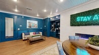 Located at 3900 Dacoma St in Houston, TX, this thoughtfully designed single-floor communal lounge offers a vibrant and functional gathering space. The room is defined by its striking deep teal wainscoting and wall paneling, which create a bold, cohesive aesthetic throughout. Warm hardwood flooring runs the length of the space, complemented by a beadboard ceiling with recessed lighting and crisp white crown molding that adds a refined finish.

A built-in nook along one wall houses a coffee and beverage station with cabinetry below and open shelving above — stocked with books, games, and décor — making it a natural hub for casual interaction. Adjacent to it, a white brick accent wall features a lush vertical moss panel with a glowing neon "AWHILE" sign and a sleek electric fireplace, adding warmth and personality to the space.

A round wooden dining table with upholstered chairs anchors the center of the room atop a patterned area rug, ideal for collaborative or social gatherings. A floral-print sofa and coffee table offer a relaxed lounge alternative nearby. Large glass doors flood the space with natural light and provide views of the outdoor area. A private, glass-enclosed phone booth pod offers a quiet retreat for focused work or calls within this otherwise open and inviting environment.