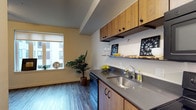 This single-floor residence offers a thoughtfully designed living space that balances functionality with a warm, contemporary aesthetic. The kitchen serves as a central highlight, featuring rich wood cabinetry with black hardware, smooth gray countertops, and a stainless steel sink. Upper cabinets provide ample storage, while open shelving adds a decorative touch, displaying glassware and accents. A built-in dishwasher contributes to the kitchen's practicality. The space opens into a living area bathed in natural light from a large window, which frames a view of the surrounding building exterior. Light-toned hardwood-style flooring runs throughout, lending a clean and cohesive look to the interior. Warm yellow walls add a welcoming brightness to the space, complemented by the greenery of an indoor potted plant that brings a natural, lively element to the room. The open layout between the kitchen and living area creates an efficient and inviting flow, making the space feel airy and functional. Overall, the design strikes a balance between modern finishes and comfortable, everyday livability.