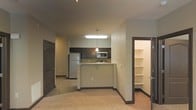 This single-floor residential unit offers a clean, functional layout designed to maximize comfort and usability. The open-concept living and kitchen area forms the heart of the space, creating a seamless flow between everyday living and cooking zones. The kitchen is outfitted with dark espresso-toned cabinetry that contrasts tastefully against the neutral beige walls and light-toned carpet flooring throughout. A peninsula-style breakfast bar with a granite-look countertop provides additional prep space and casual seating, while overhead pendant lighting adds a warm, focused glow to the kitchen area. Stainless steel appliances, including a full-size refrigerator and microwave, are neatly integrated into the cabinetry for a cohesive look. A flush ceiling light panel ensures the kitchen remains bright and well-lit. Accessible from the main living area, a generously sized walk-in closet features multiple built-in shelving units, offering excellent storage and organization potential. Dark-trimmed interior doors add a consistent design accent throughout the unit, lending a polished, modern touch to the overall aesthetic. The space presents a practical and inviting environment well-suited for comfortable day-to-day living.