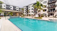 Located at 2480 First St in Fort Myers, Florida, this contemporary residential community showcases a striking resort-style outdoor amenity level designed for both relaxation and social engagement. The centerpiece is a generously sized swimming pool framed by crisp white pavers, rows of lounge chairs, and market umbrellas, all set within a symmetrical courtyard enclosed by a multi-story building facade finished in clean white with bold black-framed windows and private balconies. Towering palm trees punctuate the pool deck, reinforcing the tropical Florida ambiance throughout.

Surrounding the pool, carefully curated garden beds feature vibrant tropical plantings—bromeliads, cordylines, palms, and flowering ground cover—that add lush color and texture to the hardscaped walkways. A covered pavilion along the pool's edge provides a shaded retreat, anchored by a dramatic natural stone fireplace and furnished with comfortable seating, creating an inviting lounge atmosphere. Outdoor grilling stations with granite countertops are thoughtfully integrated into the courtyard, offering convenience for residents. The architecture maintains a modern coastal aesthetic, with clean lines, open-air corridors, and seamless indoor-outdoor connectivity that makes the most of Fort Myers' warm, sunny climate year-round.