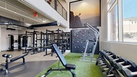 Located at 1600 W Call St in Tallahassee, FL, this modern two-story fitness facility offers a comprehensive workout environment designed to accommodate a wide range of training styles. The ground floor serves as the primary strength and conditioning zone, featuring durable rubber and turf flooring, multiple power racks with barbells, adjustable benches, and an extensive dumbbell rack bathed in natural light from large floor-to-ceiling windows. A standout "Freedom Climber" climbing wall anchors one end of the main floor, adding a dynamic functional training element. Exposed ductwork and industrial-style ceilings lend the space a contemporary athletic aesthetic, while textured accent walls provide visual warmth. An interior staircase with open metal railings connects the two floors. The upper level houses a bright, glass-walled studio room with light wood flooring, full-length mirrors, and organized storage for medicine balls, kettlebells, foam rollers, and resistance equipment — ideal for group classes or personal training. A secondary cardio area on the upper floor features rowing machines and stationary bikes overlooking the main floor below. Additional rooms provide flexible multi-use space, completing this well-appointed, high-capacity fitness center.