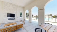 Located in Moreno Valley, CA, this covered outdoor lounge is a thoughtfully designed communal space that blends architectural elegance with casual comfort. The space is defined by a series of grand arched openings along its perimeter, which flood the interior with abundant natural light and frame picturesque views of the surrounding outdoor amenities, including a pool and landscaped grounds.

The soaring ceilings lend the lounge an airy, open feel, enhancing the sense of spaciousness throughout. A striking floor-to-ceiling stacked stone fireplace serves as the room's focal point, integrated with a wall-mounted television, making it an ideal setting for both relaxed gatherings and entertainment.

Underfoot, large-format light concrete tiles extend across the entire space, contributing to the clean, modern aesthetic. The lounge is furnished with teak-framed outdoor seating, including sofas, lounge chairs, and a coffee table, all upholstered in neutral grey cushions and arranged over a patterned area rug to create an inviting conversation area.

A dining table with chairs is positioned toward one end, accompanied by easy access through glass-panel doors that connect the space to adjacent indoor amenities. The overall ambiance is bright, welcoming, and resort-inspired, making this lounge a standout communal feature of the property.
