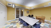 This single-floor community room is a welcoming, well-appointed space designed to accommodate a variety of group activities and gatherings. The room is defined by warm yellow walls that lend an inviting, energetic atmosphere, complemented by a colorful multi-toned checkerboard vinyl tile floor featuring shades of blue, gold, grey, and white.

Multiple large rectangular tables with navy blue chairs are arranged throughout the space, offering flexible configurations for dining, meetings, or collaborative activities. Additional natural wood tables and chairs provide supplementary seating options. Overhead, linear pendant light fixtures provide ample illumination across the room.

Natural light filters in through windows dressed with patterned valances, offering views of the surrounding neighborhood. Framed artwork adorns the walls, adding a touch of character to the space.

A built-in kitchenette area is equipped with dark wood cabinetry — both upper and lower — a stainless steel sink with a modern gooseneck faucet, and light-toned countertops, making the room well-suited for catered events or light food preparation.

A set of glass-paned double doors connects to an adjacent area, enhancing the sense of openness and allowing for easy flow between spaces. Overall, the room combines functionality with a cheerful, community-oriented aesthetic.