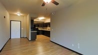 This single-story space showcases an open-concept design that seamlessly connects the living area to the kitchen. Light hardwood flooring extends throughout, creating a warm and cohesive aesthetic. The living area is equipped with a ceiling fan, adding both function and a casual comfort to the space.

The kitchen is thoughtfully designed with rich, dark wood cabinetry that extends to the upper walls, offering generous storage. Black appliances, including a range and overhead microwave, complement the cabinetry for a coordinated look. A peninsula provides additional counter space and serves as a natural divider between the kitchen and living areas. The kitchen also features a sink positioned along the back counter, tile backsplash detailing, and overhead flush-mount lighting.

Neutral-toned walls throughout the space provide a versatile backdrop, while black baseboards offer a subtle contrast to the light flooring. Multiple electrical outlets are conveniently placed along the walls. The overall ambiance is clean, modern, and functional, with an efficient layout that maximizes the available space.