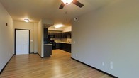 This single-story space showcases an open-concept design that seamlessly connects the living area to the kitchen. Light hardwood flooring extends throughout, creating a warm and cohesive aesthetic. The living area is equipped with a ceiling fan, adding both function and a casual comfort to the space.

The kitchen is thoughtfully designed with rich, dark wood cabinetry that extends to the upper walls, offering generous storage. Black appliances, including a range and overhead microwave, complement the cabinetry for a coordinated look. A peninsula provides additional counter space and serves as a natural divider between the kitchen and living areas. The kitchen also features a sink positioned along the back counter, tile backsplash detailing, and overhead flush-mount lighting.

Neutral-toned walls throughout the space provide a versatile backdrop, while black baseboards offer a subtle contrast to the light flooring. Multiple electrical outlets are conveniently placed along the walls. The overall ambiance is clean, modern, and functional, with an efficient layout that maximizes the available space.
