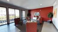 This single-floor communal space makes a striking first impression with its bold design language, combining deep red accent walls, warm gray tones, and contemporary furnishings throughout. The main lounge area is anchored by large floor-to-ceiling windows that flood the space with natural light and offer views of the surrounding neighborhood. Comfortable upholstered armchairs with red accent pillows are arranged around a round white coffee table, creating an inviting setting for relaxation or conversation. A bold, patterned bench adds a lively focal point to the seating arrangement, while a sleek white console table with a decorative mirror is set against the signature red wall. Adjacent to the lounge, a dedicated media room features wall-mounted speakers, built-in cabinetry with frosted glass panels, and a large flat-screen television — all complemented by striking red accent chairs that reinforce the space's vibrant color palette. The corridor area connects the spaces seamlessly, adorned with bold abstract artwork in warm orange and red tones. A centrally located elevator framed by deep red wall panels provides vertical access, while a staircase with modern metal railings adds architectural interest. The overall ambiance is energetic yet comfortable, blending modern design with functional communal living.