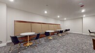 Located at 2 Walter Ave in Highland Park, NJ, this single-floor communal space is thoughtfully designed to accommodate both casual gatherings and focused work. The main area features a defining built-in banquette that runs along one wall, upholstered in neutral tan acoustic panels framed by rich wood trim, providing both comfort and sound absorption. Paired with round white pedestal tables and contemporary navy chairs with gold-toned metal legs, the seating arrangement offers an inviting setting for small group interactions.

The open layout extends into a lounge area furnished with plush upholstered armchairs and accent side tables, creating a relaxed environment ideal for informal meetings or casual conversation. Patterned grey carpeting flows throughout, adding texture and warmth to the clean, neutral-toned walls. Recessed lighting illuminates the entire space evenly, contributing to a clean and polished atmosphere.

A secondary alcove area provides additional seating with a round table and chairs, offering a more intimate setting for one-on-one conversations or quiet work. The space is accessible via double entry doors and includes a separate private door, ensuring flexibility in use. Overall, the space strikes a balance between professional functionality and modern comfort.