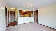 This single-floor residence offers a functional and straightforward layout designed for comfortable everyday living. The open-concept living area features wall-to-wall carpeting that lends a warm, understated feel to the space. A defining architectural feature is the half-wall pass-through that connects the living area to the kitchen, creating a sense of openness while maintaining a defined cooking zone. The kitchen is equipped with rich, dark wood cabinetry providing ample storage above the countertops, complemented by a refrigerator and range. Track lighting runs along the kitchen ceiling, delivering focused illumination across the workspace, while a globe pendant light adds a subtle decorative touch. The neutral-toned walls and textured ceiling keep the aesthetic clean and unobtrusive. Dark wood trim borders the baseboards and door frames throughout, tying the interior palette together cohesively. Two doorways branch off from the main living space, offering access to additional rooms and lending the layout a practical flow. The entry area features hardwood-style flooring, providing a durable transition point into the carpeted interior. Overall, the space presents a classic, no-frills residential design with an efficient use of its single-floor footprint.