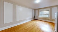 This single-floor unit offers a thoughtfully arranged layout comprising a living room, bathroom, closet, and connecting hallway. The living room serves as the centerpiece of the space, showcasing warm-toned hardwood floors that run throughout and neutral, greige-painted walls that lend a calm, versatile backdrop. A cast-iron radiator positioned beneath a window provides both character and functional heating, while the window allows natural light to filter in. The walls feature a series of recessed white panels, adding subtle architectural interest and a clean, structured aesthetic. A dedicated closet with a built-in shelf and hanging rod provides practical storage. The hallway connects the main living area to the bathroom efficiently, maintaining a smooth flow throughout the unit. The bathroom is finished with floor-to-ceiling light beige ceramic tile surrounding a full soaking tub, complemented by a frosted glass window that introduces soft, diffused natural light while maintaining privacy. White trim and baseboards throughout the unit tie the design together, reinforcing a crisp, classic finish. The overall ambiance is understated and functional, with traditional details offering a timeless quality well-suited to everyday living.