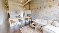 Located at 1277 West 6th Street in Cleveland's vibrant Ohio City neighborhood, this 2-bedroom, 1-bathroom loft-style residence seamlessly blends historic industrial character with contemporary design. The open-concept kitchen and living area is the heart of the unit, showcasing dramatically tall ceilings, original exposed brick walls, and dark hardwood floors. The kitchen is outfitted with white shaker cabinetry, quartz countertops, stainless steel appliances including a French door refrigerator, a smooth-top range with hood, and a peninsula with bar seating. The living area features large arched windows with built-in window seating, flooding the space with natural light and offering views of the surrounding cityscape. A long, light-filled hallway with pendant lighting leads to the two bedrooms, both offering generous ceiling heights and hardwood flooring. The bathroom is elegantly appointed with a dual-sink white vanity, quartz countertops, matte black fixtures, a large frameless mirror with industrial-style lighting, and a glass-enclosed marble-tiled shower. The combination of raw architectural elements and polished modern finishes gives this space a distinctive, upscale urban character.