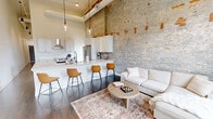 Located at 1277 West 6th Street in Cleveland's vibrant Ohio City neighborhood, this 2-bedroom, 1-bathroom loft-style residence seamlessly blends historic industrial character with contemporary design. The open-concept kitchen and living area is the heart of the unit, showcasing dramatically tall ceilings, original exposed brick walls, and dark hardwood floors. The kitchen is outfitted with white shaker cabinetry, quartz countertops, stainless steel appliances including a French door refrigerator, a smooth-top range with hood, and a peninsula with bar seating. The living area features large arched windows with built-in window seating, flooding the space with natural light and offering views of the surrounding cityscape. A long, light-filled hallway with pendant lighting leads to the two bedrooms, both offering generous ceiling heights and hardwood flooring. The bathroom is elegantly appointed with a dual-sink white vanity, quartz countertops, matte black fixtures, a large frameless mirror with industrial-style lighting, and a glass-enclosed marble-tiled shower. The combination of raw architectural elements and polished modern finishes gives this space a distinctive, upscale urban character.