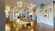 This expansive single-floor amenity space offers a thoughtfully designed interior that blends upscale residential warmth with functional communal living. Rich, wide-plank hardwood flooring flows continuously throughout, anchoring the open layout and connecting each distinct zone seamlessly.

Signature whitewashed stone columns rise from floor to ceiling across the space, serving as striking architectural focal points that define areas without interrupting the open sightlines. Coffered and tray ceilings add dimension overhead, while layered lighting — including a dramatic brass ring chandelier and a cascading crystal fixture — creates an inviting and polished ambiance.

The main lounge area features multiple curated seating arrangements, including accent chairs in bold yellows and blues centered around coffee tables atop soft area rugs. Floor-to-ceiling glass windows and glazed entry doors flood the space with natural light and provide views of mature trees outside, reinforcing a connection to the outdoors.

A dedicated conference or co-working area is outfitted with a long white communal table flanked by upholstered chairs in soft blues and grays, complemented by dark built-in shelving lined with books and decorative objects. Artwork throughout adds color and character.

A light-filled corridor with glass-paneled partitions leads to additional private rooms, restrooms, and service areas, completing this well-appointed and versatile communal environment.