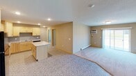 This single-floor unit is designed with a practical, open-concept layout that seamlessly connects the kitchen and living area, creating a spacious and versatile environment. The kitchen is well-appointed with light maple cabinetry, granite-patterned countertops, a central island for additional prep space, black appliances including an electric range, microwave, and refrigerator, and a double stainless steel sink. Recessed lighting illuminates the space, and a distinctive patterned tile floor adds visual interest.

The living area flows naturally from the kitchen, featuring neutral-toned carpet, a baseboard heating system, and a wall-mounted air conditioning unit alongside a window fitted with blinds, providing natural light and outdoor views. Two carpeted bedrooms follow a similar design language, each offering a window with blinds, baseboard heating, and a textured ceiling with flush-mount lighting.

The unit includes two bathrooms. The primary bathroom features a walk-in shower, a light maple vanity with a white countertop, and tile flooring. The secondary bathroom mirrors this style, offering a vanity with a white countertop, a lighted mirror, and matching tile flooring.