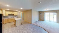 This single-floor unit is designed with a practical, open-concept layout that seamlessly connects the kitchen and living area, creating a spacious and versatile environment. The kitchen is well-appointed with light maple cabinetry, granite-patterned countertops, a central island for additional prep space, black appliances including an electric range, microwave, and refrigerator, and a double stainless steel sink. Recessed lighting illuminates the space, and a distinctive patterned tile floor adds visual interest.

The living area flows naturally from the kitchen, featuring neutral-toned carpet, a baseboard heating system, and a wall-mounted air conditioning unit alongside a window fitted with blinds, providing natural light and outdoor views. Two carpeted bedrooms follow a similar design language, each offering a window with blinds, baseboard heating, and a textured ceiling with flush-mount lighting.

The unit includes two bathrooms. The primary bathroom features a walk-in shower, a light maple vanity with a white countertop, and tile flooring. The secondary bathroom mirrors this style, offering a vanity with a white countertop, a lighted mirror, and matching tile flooring.